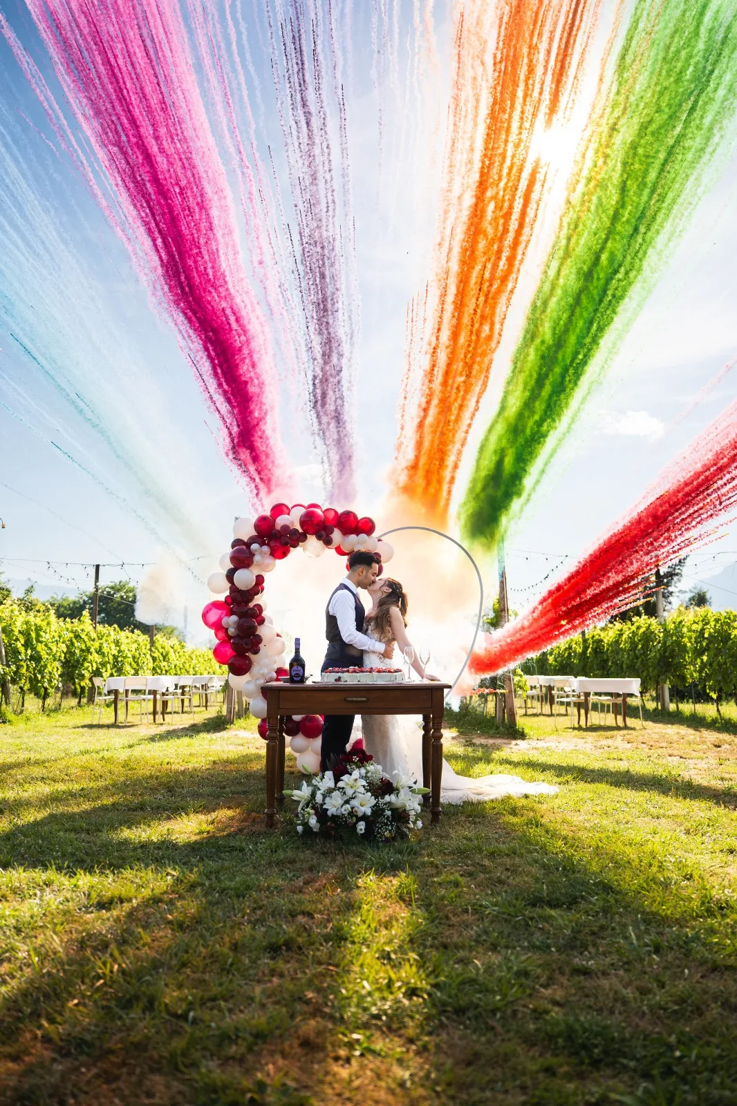 Gli sposi si baciano in vigna sotto i fumogeni colorati, con un arco di palloncini e il tavolo della torta